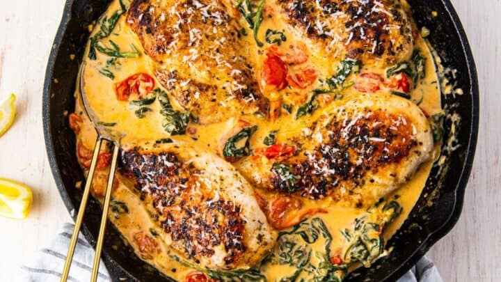 A cast iron skillet with four seared chicken breasts in a creamy sauce with spinach and cherry tomatoes, garnished with grated cheese. A cloth napkin and lemon wedges are nearby—perfect for budget friendly dinner recipes.