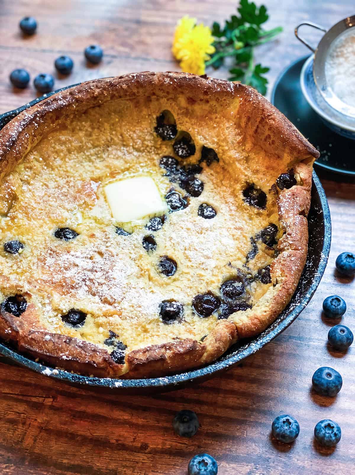 A Dutch baby pancake with blueberries, featuring a golden puff and a pat of butter in the center, is surrounded by fresh blueberries on a rustic wooden table.