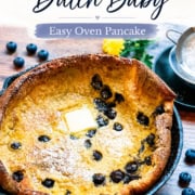 A classic Dutch baby pancake, baked to golden perfection in a skillet, is topped with a pat of butter and surrounded by fresh blueberries and a bright lemon slice.