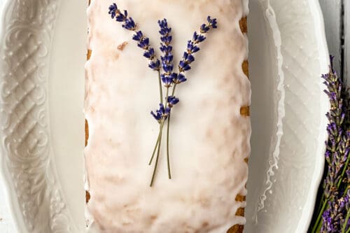 A rectangular glazed lavender tea bread topped with sprigs of lavender sits on an ornate white platter. Beside the platter is a small bouquet of lavender tied with a purple and green checkered ribbon, all placed on a white wooden surface.