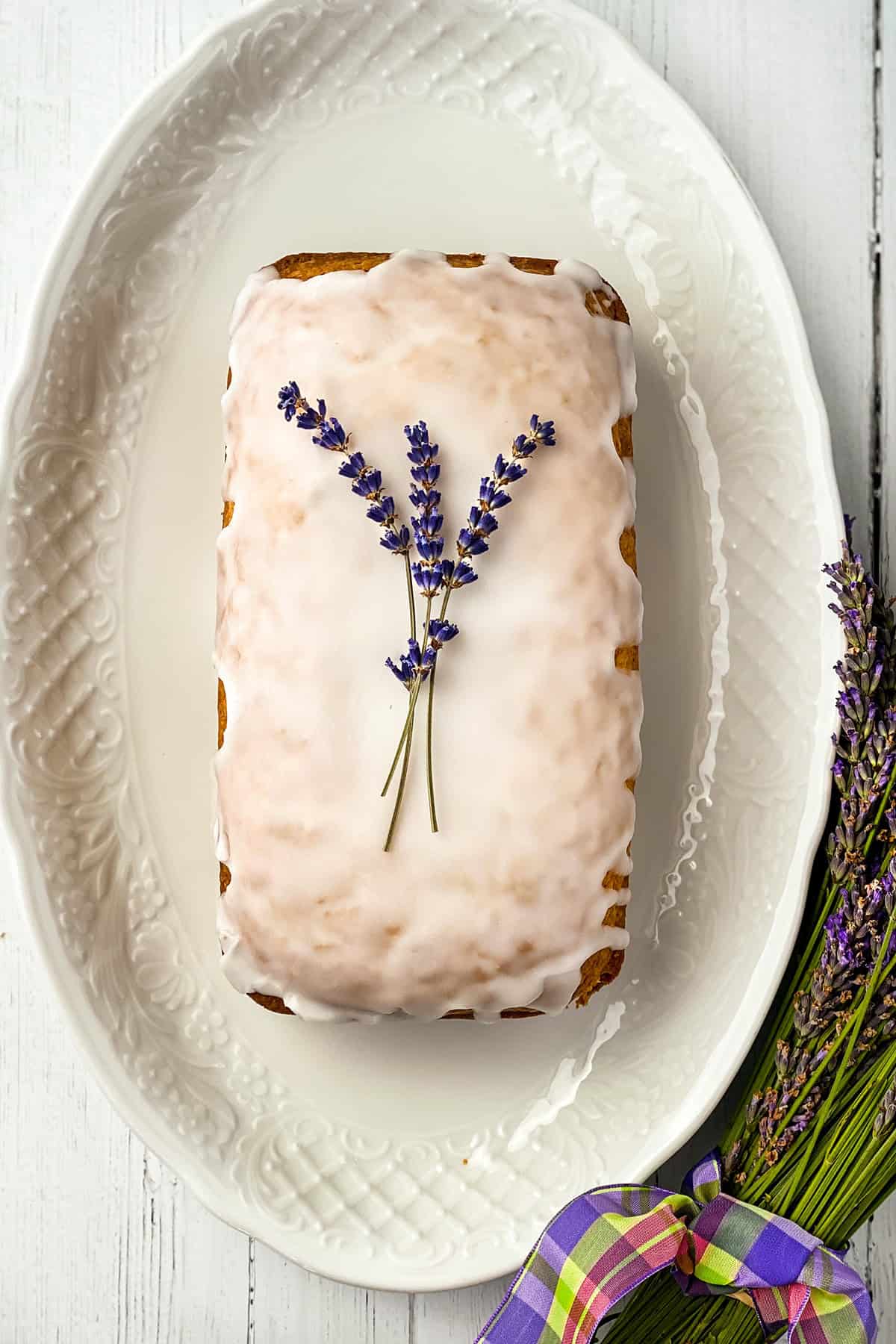 A rectangular lavender tea bread loaf with white icing, topped with two sprigs of lavender, is placed on a white oval platter next to a small bouquet of lavender tied with a plaid ribbon.