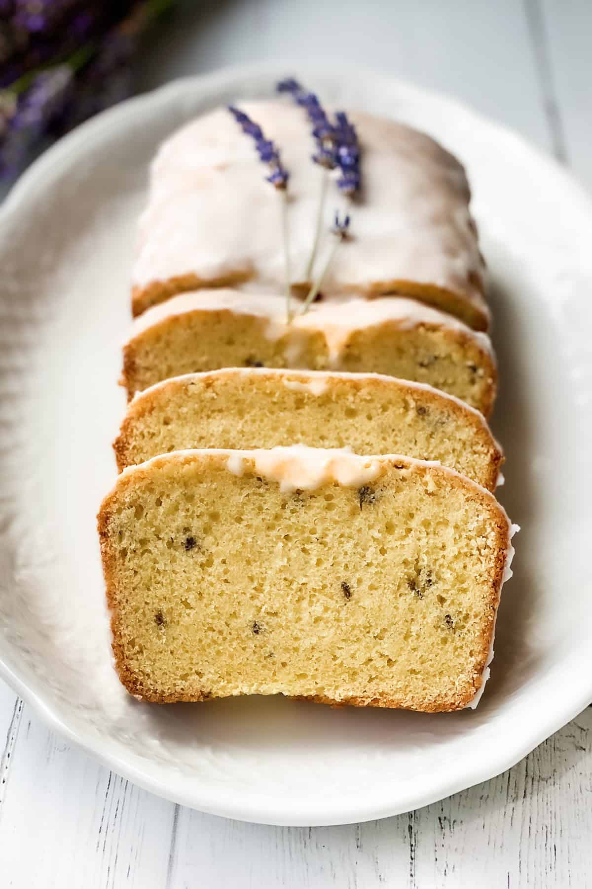 A loaf of lavender tea bread with icing, partially sliced and arranged on a white plate, garnished with sprigs of lavender.