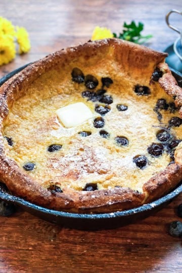 A delicious dutch baby pancake in a cast iron skillet, filled with juicy blueberries and topped with a pat of butter and a dusting of powdered sugar, with fresh blueberries scattered around.