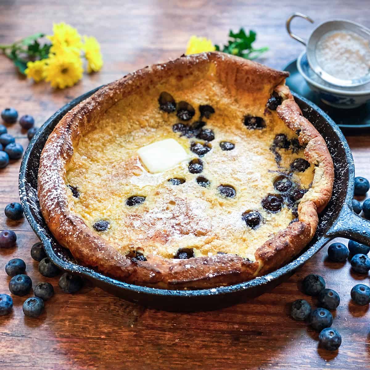 A delicious dutch baby pancake in a cast iron skillet, filled with juicy blueberries and topped with a pat of butter and a dusting of powdered sugar, with fresh blueberries scattered around.