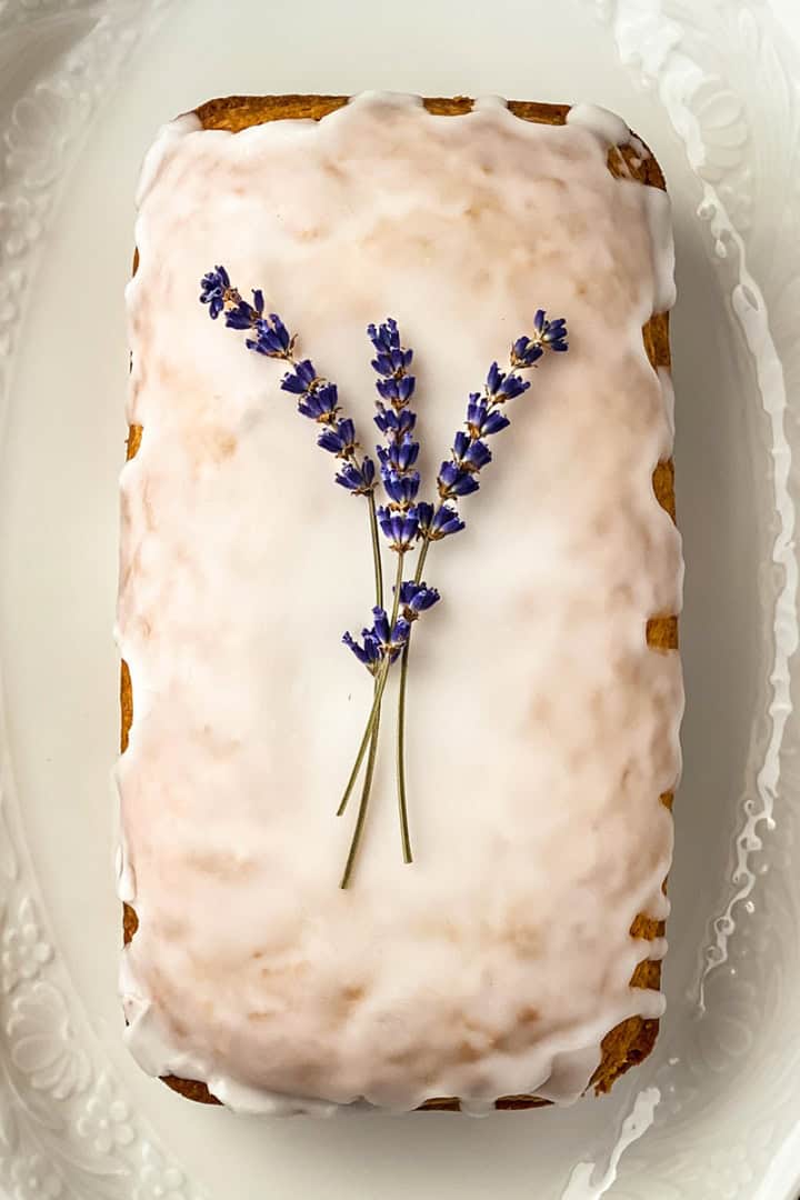 A rectangular lavender tea bread loaf with white icing, garnished with two sprigs of lavender, displayed on a white, textured oval plate.