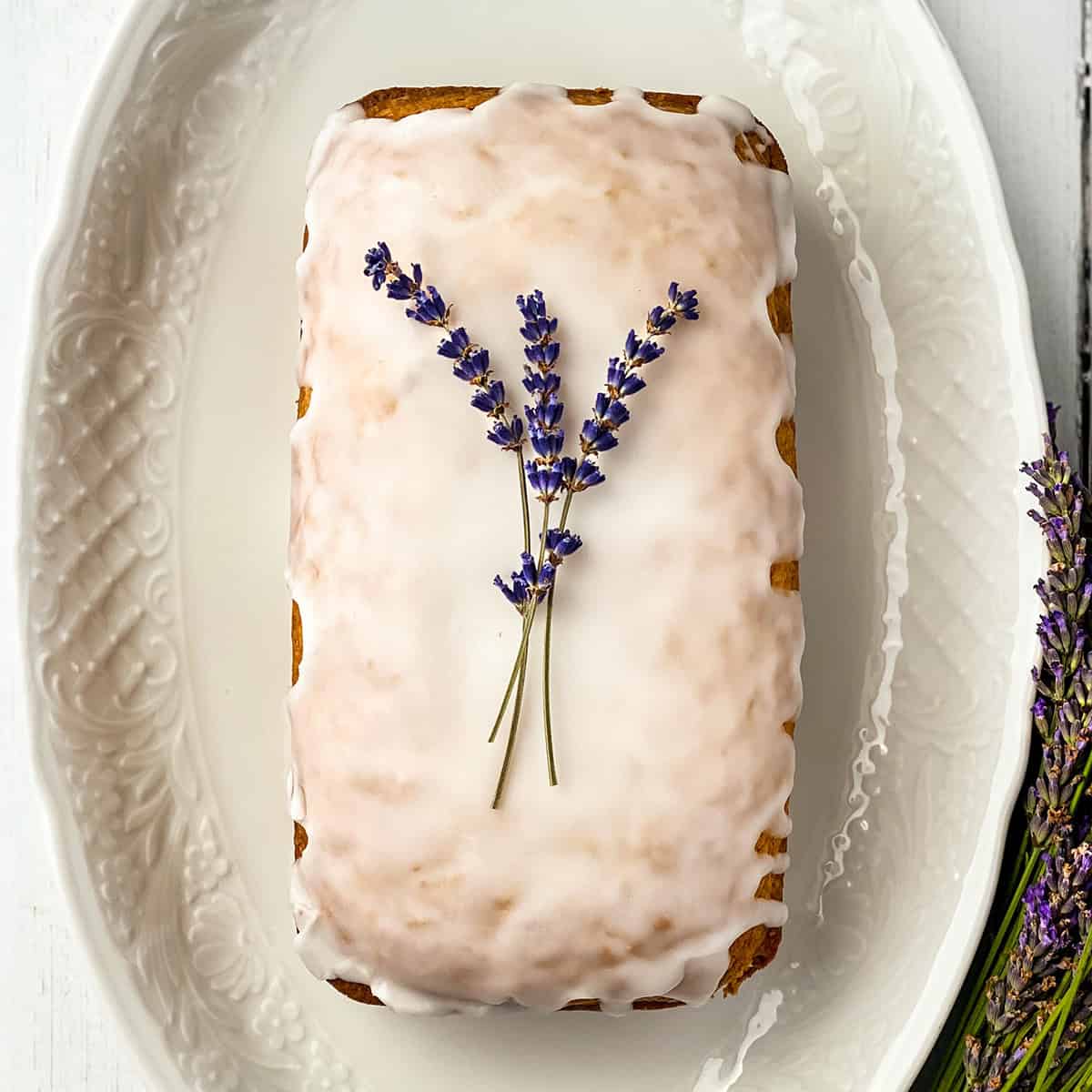 A rectangular lavender tea bread loaf with white icing, garnished with two sprigs of lavender, displayed on a white, textured oval plate.