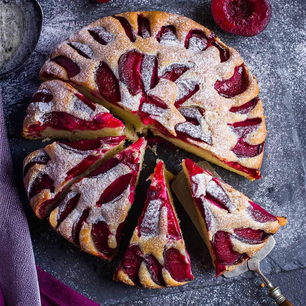 Top view of partially sliced plum cake with a silver server and visible plum slices.
