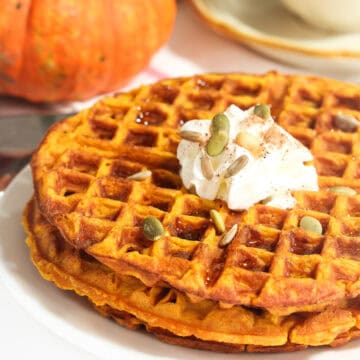 Stacked pumpkin waffles on a plate topped with whipped cream and pumpkin seeds.