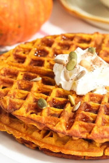 Stacked pumpkin waffles on a plate topped with whipped cream and pumpkin seeds.