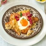 A plate with crispy sheet pan breakfast hash browns, bacon strips, and a sunny-side-up egg, garnished with chives, served on a decorative brown and white plate.