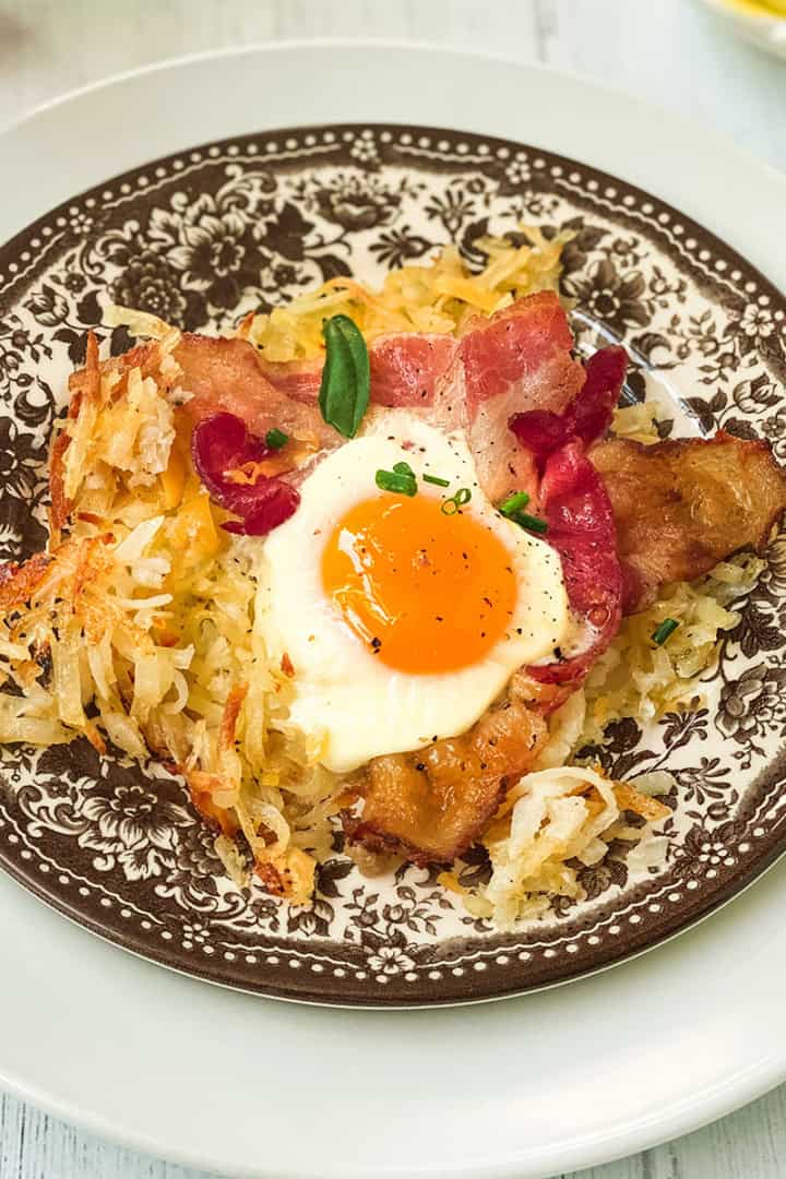 A plate with crispy sheet pan breakfast hash browns, bacon strips, and a sunny-side-up egg, garnished with chives, served on a decorative brown and white plate.