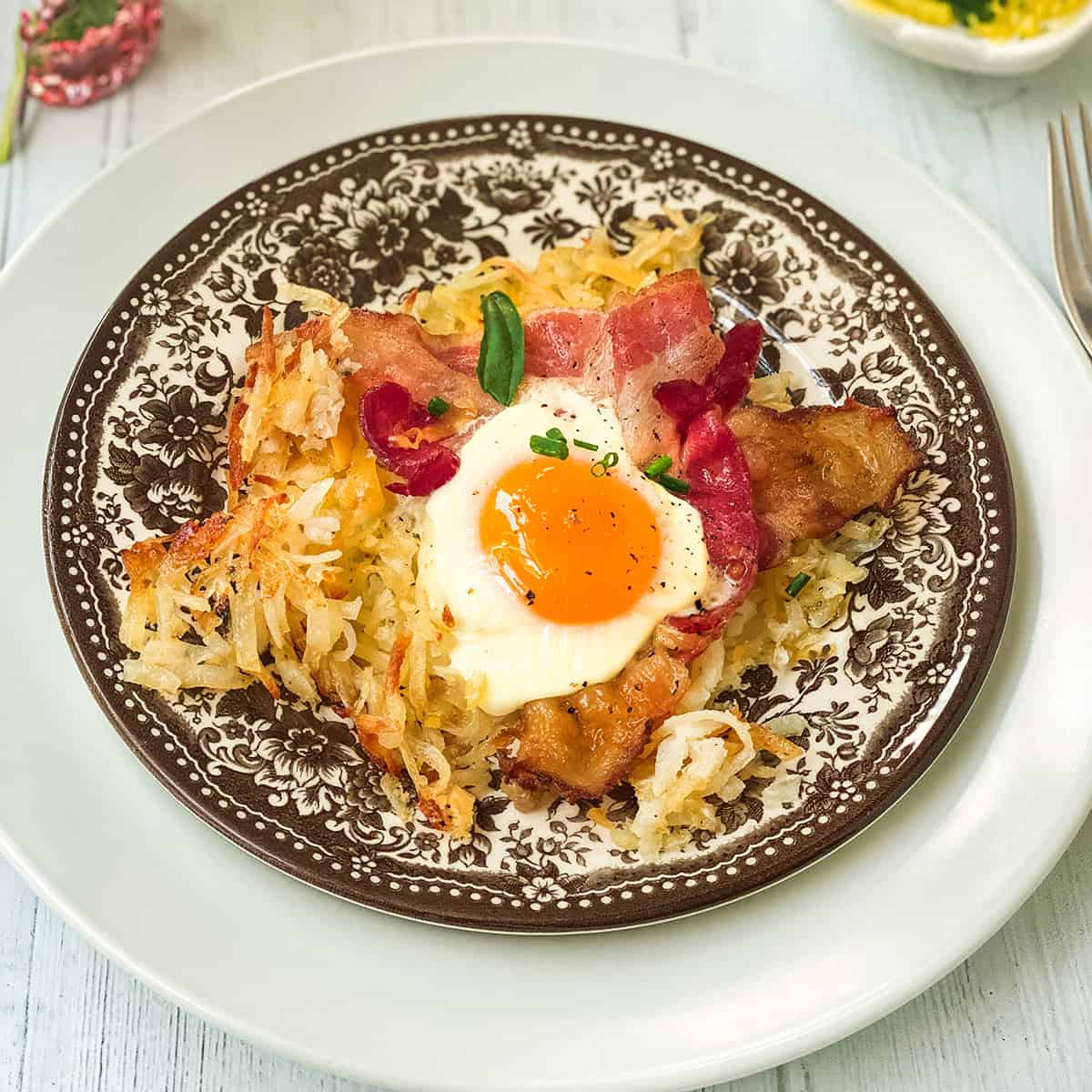 A plate with crispy sheet pan breakfast hash browns, bacon strips, and a sunny-side-up egg, garnished with chives, served on a decorative brown and white plate.