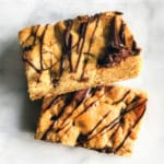 Two rectangular Chocolate Chip Cookie Bars with chocolate drizzle, stacked on a white surface.