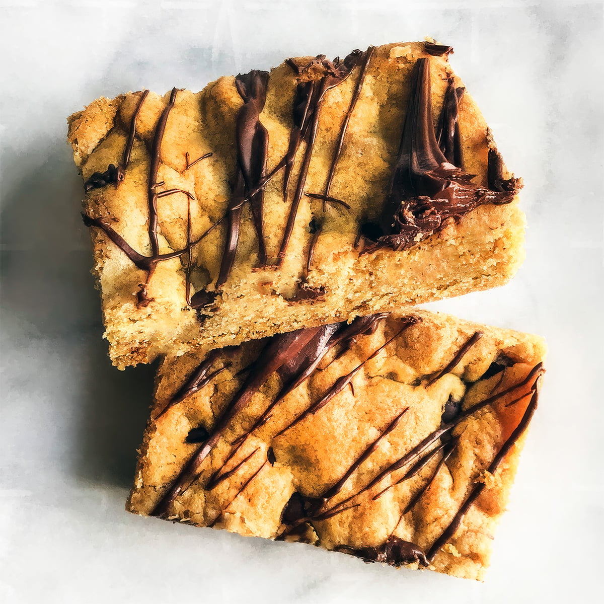 Two rectangular Chocolate Chip Cookie Bars with chocolate drizzle, stacked on a white surface.