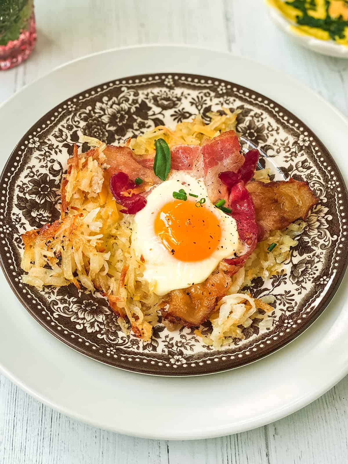 A plate with hash browns, strips of bacon, and a sunny-side-up egg on top, garnished with fresh herbs-this sheet pan breakfast is served on a patterned brown and white dish.