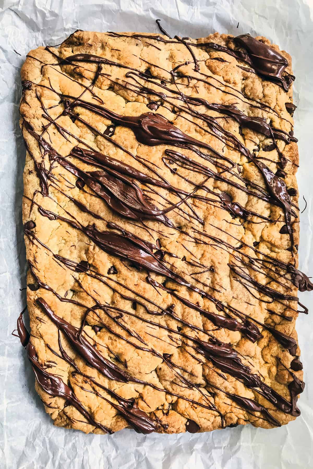 A large rectangular Chocolate Chip Cookie Bars slab, drizzled with melted chocolate, rests on crinkled parchment paper.