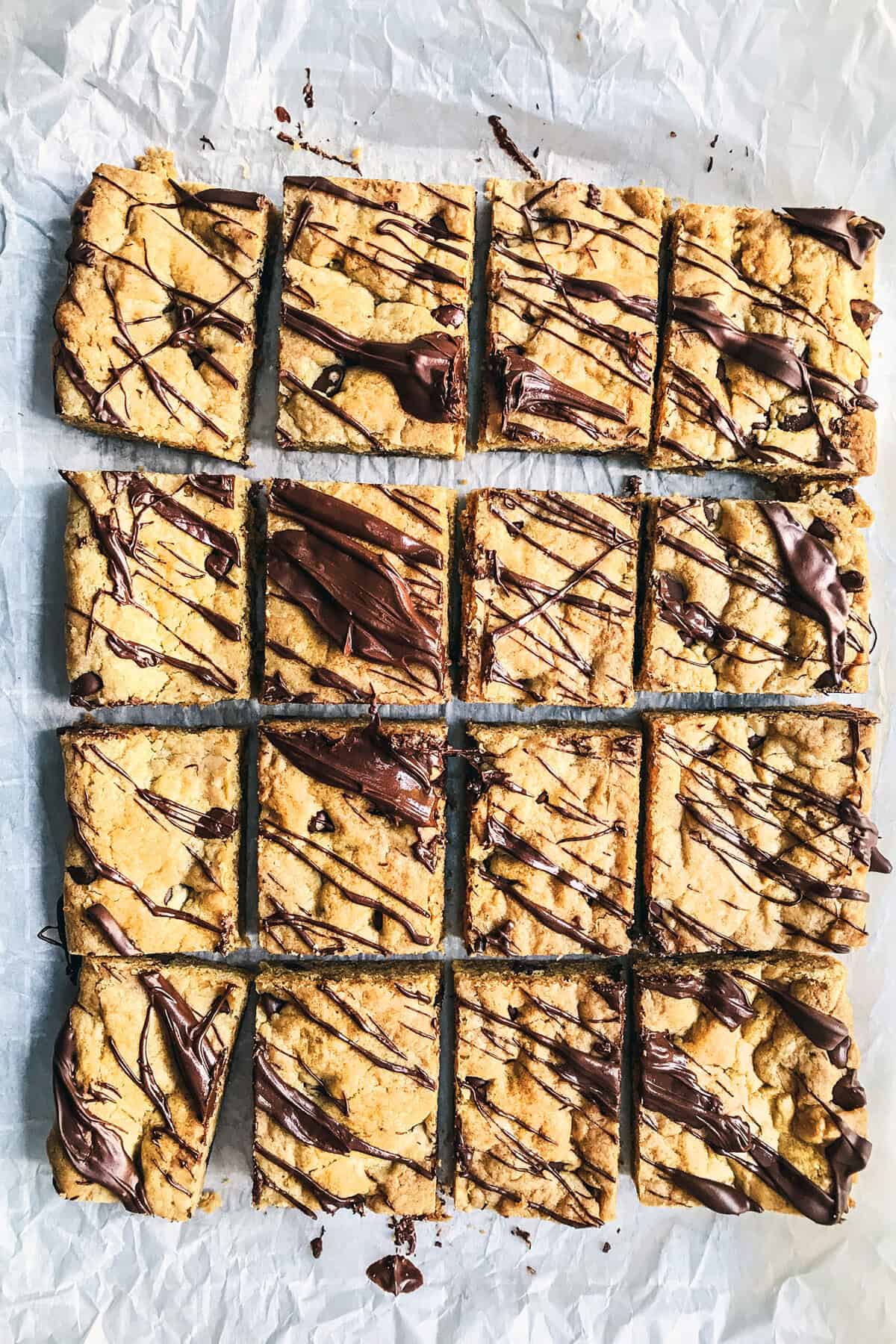 Sixteen Chocolate Chip Cookie Bars arranged in a grid on parchment paper, each topped with a drizzle of melted chocolate. The bars have a golden-brown color and visible chocolate chips.