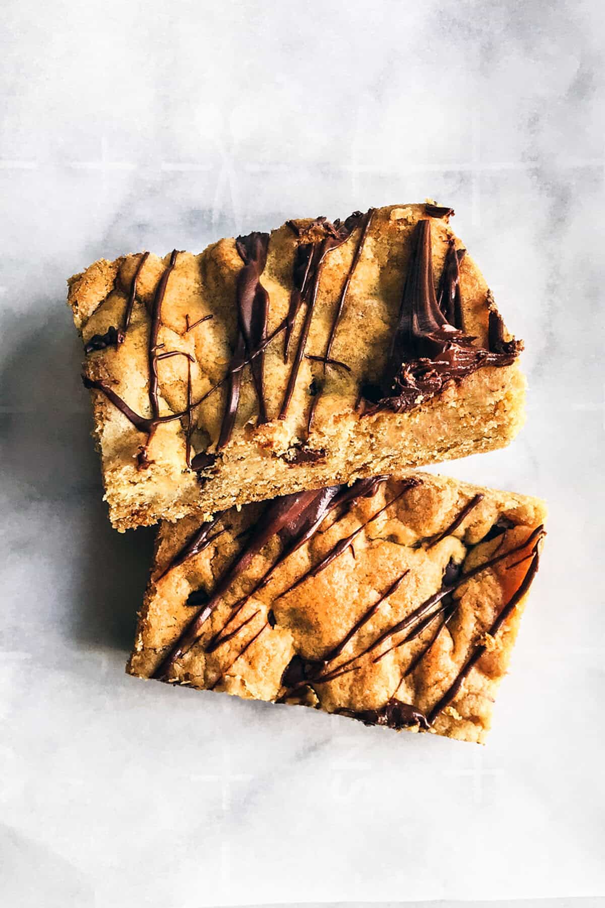 Two rectangular chocolate chip cookie bars stacked on each other, drizzled with chocolate on top, placed on a white parchment paper background.