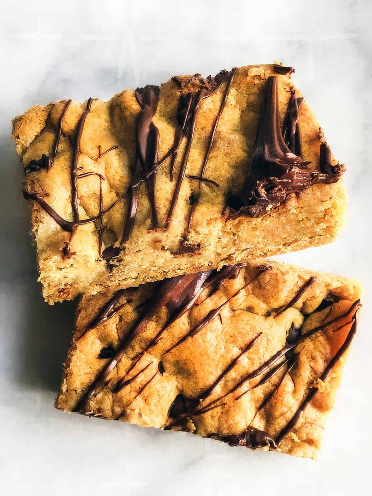 Two rectangular Chocolate Chip Cookie Bars topped with drizzled melted chocolate, stacked on a light-colored surface.