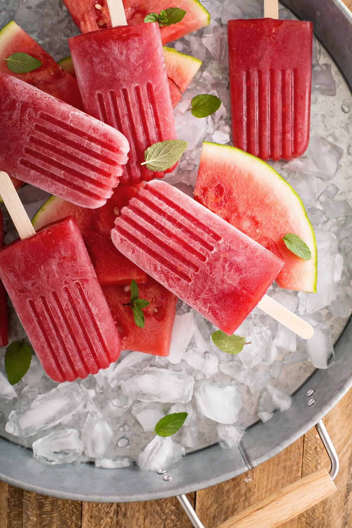 Watermelon Mint Popsicles and fresh watermelon slices arranged on ice in a metal tray, with mint leaves scattered on top.