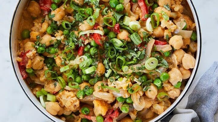 A pan filled with a chickpea, pea, and vegetable stir-fry garnished with chopped herbs rests on a blue and white striped towel—perfect for summer 15 minute dinners.