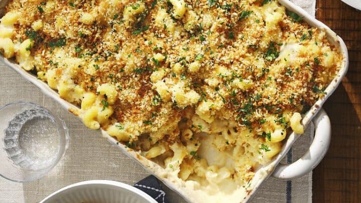 A casserole dish of baked macaroni and cheese with a breadcrumb topping sits on a table, next to two plates—one with a serving and fork—making it a classic choice for budget friendly dinner recipes.