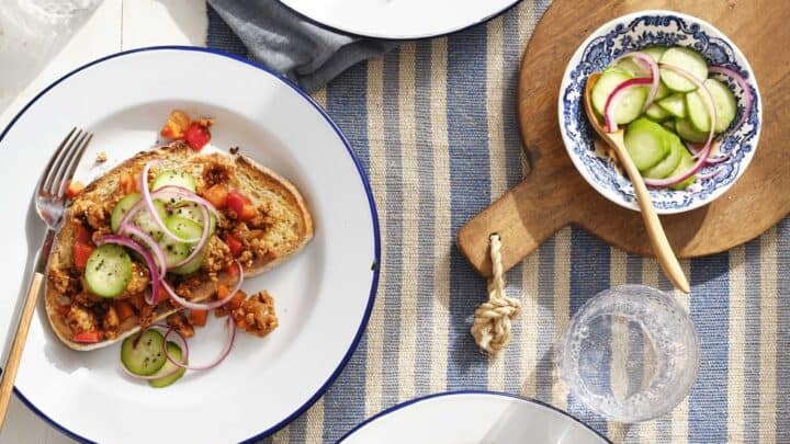 Three plates with open-faced sandwiches topped with a vegetable and grain mixture, sliced cucumber, and red onion; a bowl of cucumber salad and utensils are also on the table—perfect for budget friendly dinner recipes.