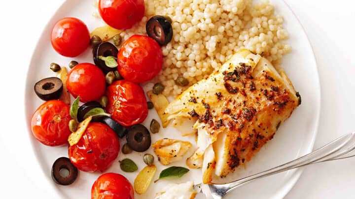 A plate with baked fish, pearl couscous, cherry tomatoes, black olives, and herbs—perfect for late summer dinners. A fork rests on the plate with a piece of fish.