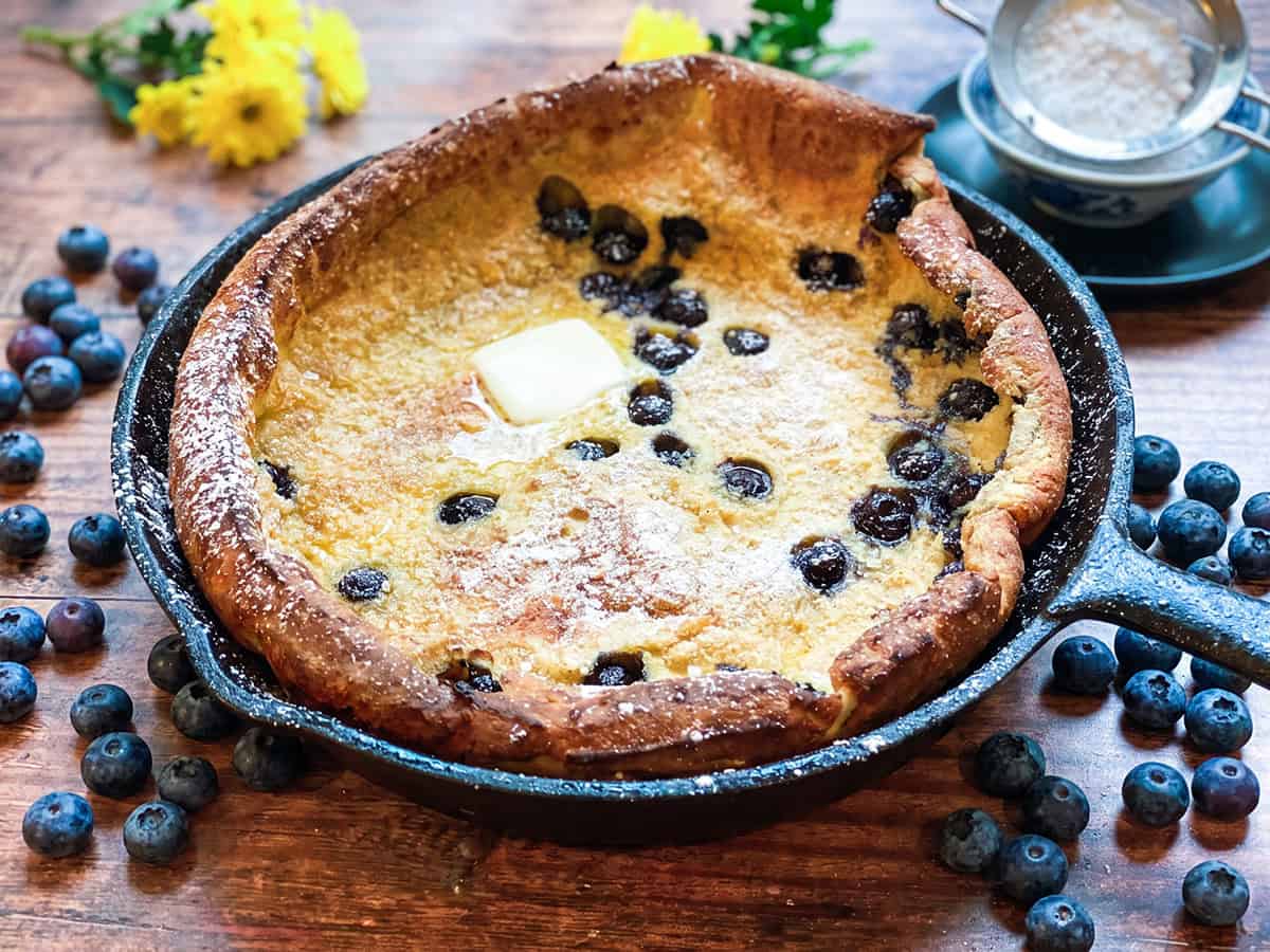 A classic dutch baby pancake with juicy blueberries baked in a cast iron skillet, topped with a pat of butter and a dusting of powdered sugar.