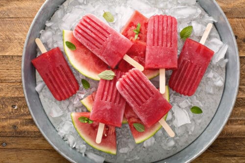 A round metal tray filled with ice, watermelon slices, and several bright red Watermelon Mint Popsicles with wooden sticks, garnished with fresh mint leaves. The tray sits on a wooden surface.