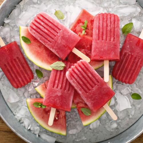 A round metal tray filled with ice, watermelon slices, and several bright red Watermelon Mint Popsicles with wooden sticks, garnished with fresh mint leaves. The tray sits on a wooden surface.