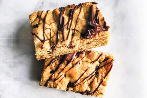 Two rectangular chocolate chip cookie bars, drizzled with melted chocolate, are stacked on a white marble surface for an irresistible treat.