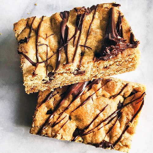 Two rectangular chocolate chip cookie bars, drizzled with melted chocolate, are stacked on a white marble surface for an irresistible treat.