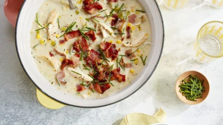 A pot and a bowl of creamy soup topped with crispy bacon and herbs, alongside empty bowls, spoons, glasses, and a small dish of chopped chives—perfect for those seeking budget friendly dinner recipes.