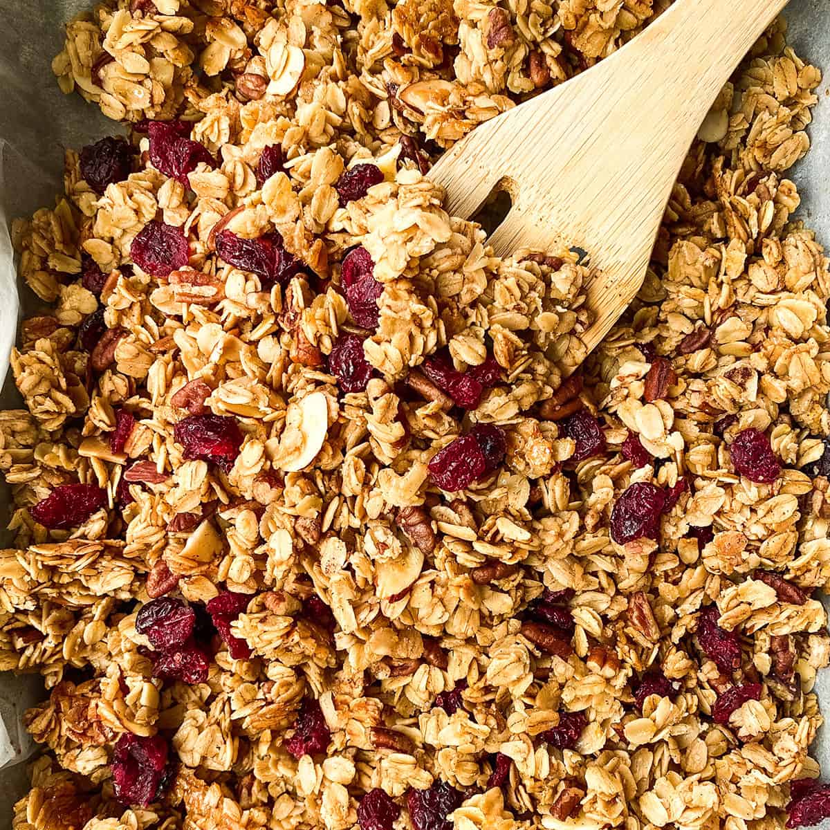 Baked healthy granola with dried fruit in a sheet pan with a wooden spoon, cooling.