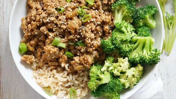 A bowl of brown rice topped with cooked ground meat, sesame seeds, chopped green onions, and steamed broccoli—perfect for summer 15 minute dinners—with lime wedges, cilantro, and chopsticks nearby on a wooden surface.
