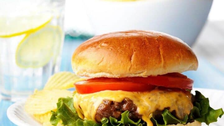 A classic cheeseburger with lettuce and tomato on a bun, served with potato chips on a white plate—perfect for celebrating Labor Day Week. A glass of lemon water and extra chips complete the festive meal in the background.