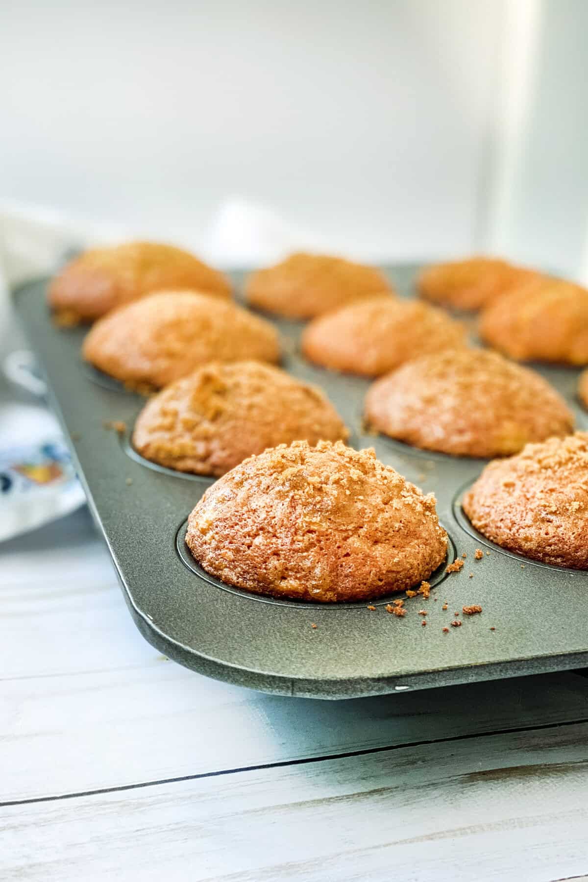 A close-up of freshly baked banana muffins in a metal tray. The muffins are golden brown with a crumbly topping, showcasing a delightful banana muffin recipe, placed on a light-colored wooden surface.