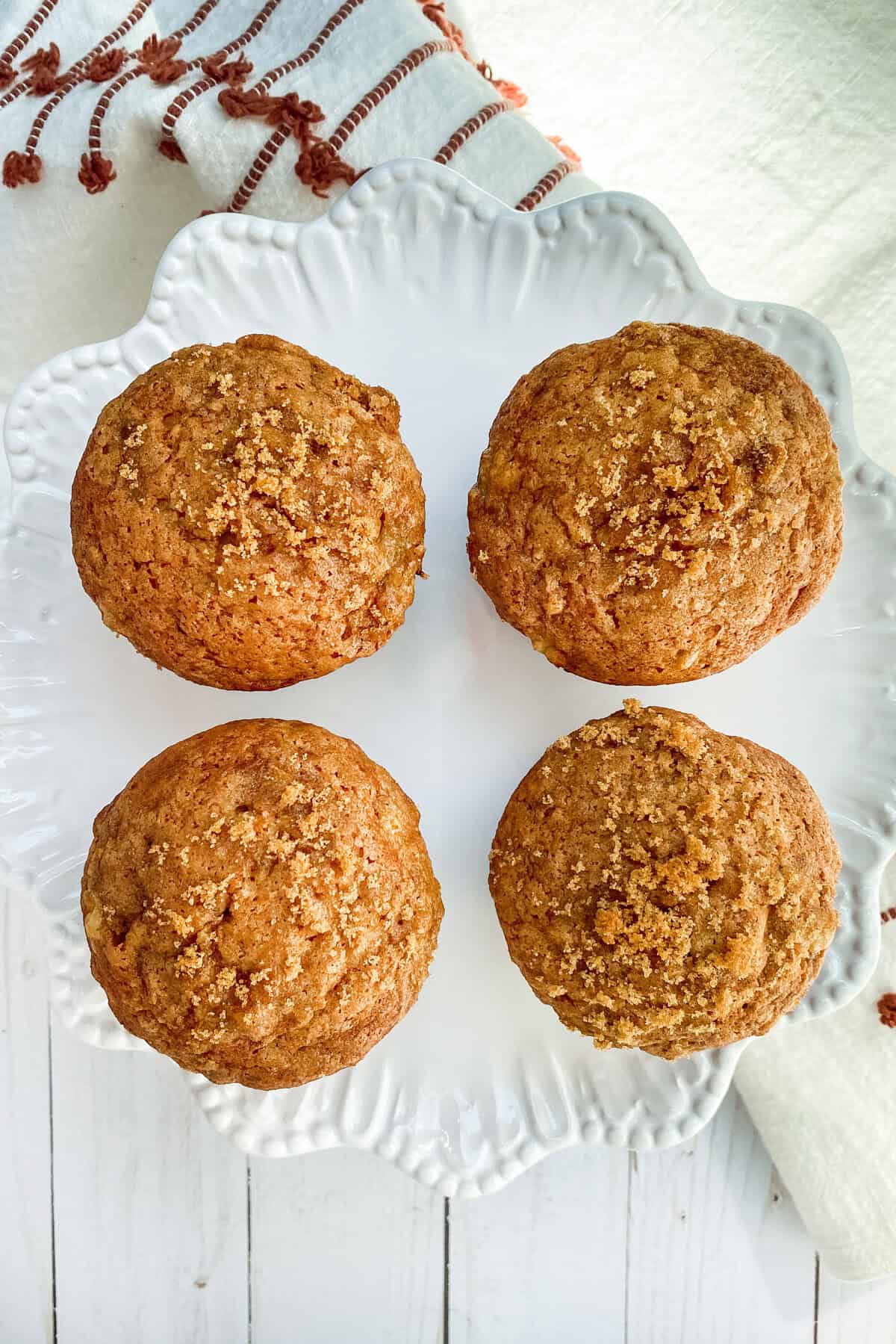 Four freshly baked banana muffins sit on a decorative white plate, resting on a white wooden surface. A red-striped cloth is partially visible in the top left corner. The muffins have a light brown, crumbly texture, perfect for those seeking an irresistible banana muffin recipe.