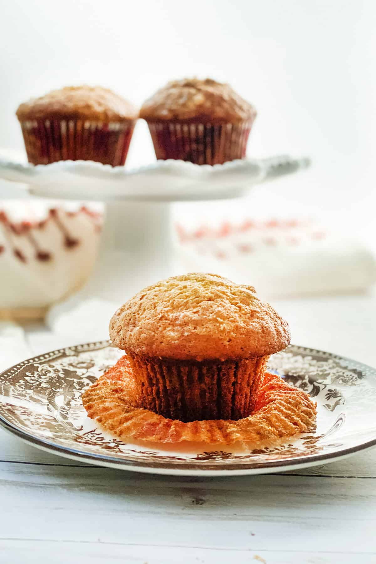 A close-up of a freshly baked banana muffin on a decorative plate, with its wrapper partially peeled. In the background, more muffins sit on a cake stand, softly out of focus, against a bright, airy backdrop—perfectly presenting this delightful banana muffin recipe.