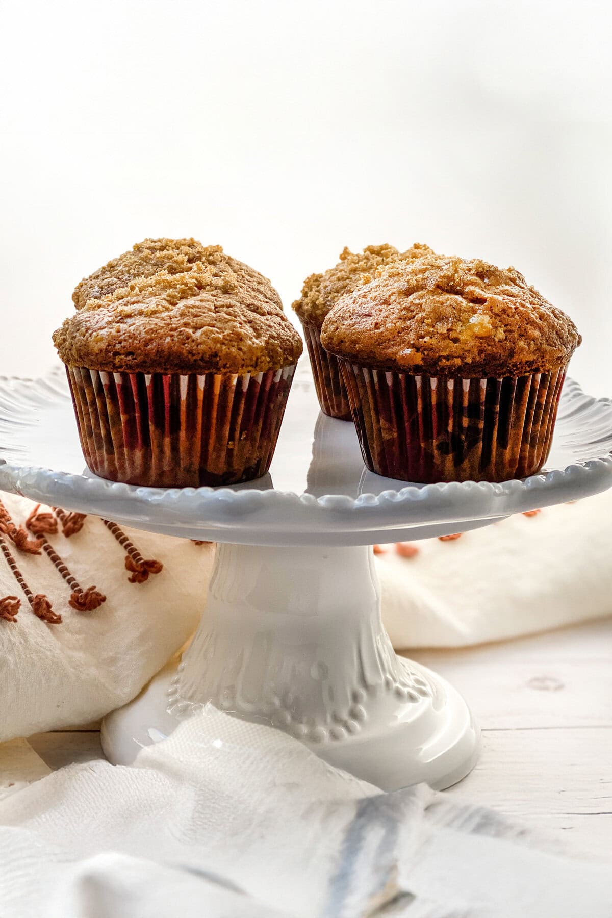 Three banana muffins with golden-brown tops sit on a white cake stand. The backdrop includes a white cloth with brown embroidery and a white wooden surface, crafting a cozy, homemade atmosphere that whispers of cherished recipes.