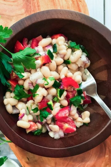A wooden bowl filled with White Bean Salad, featuring white beans, chopped tomatoes, onions, parsley, and a metal spoon.