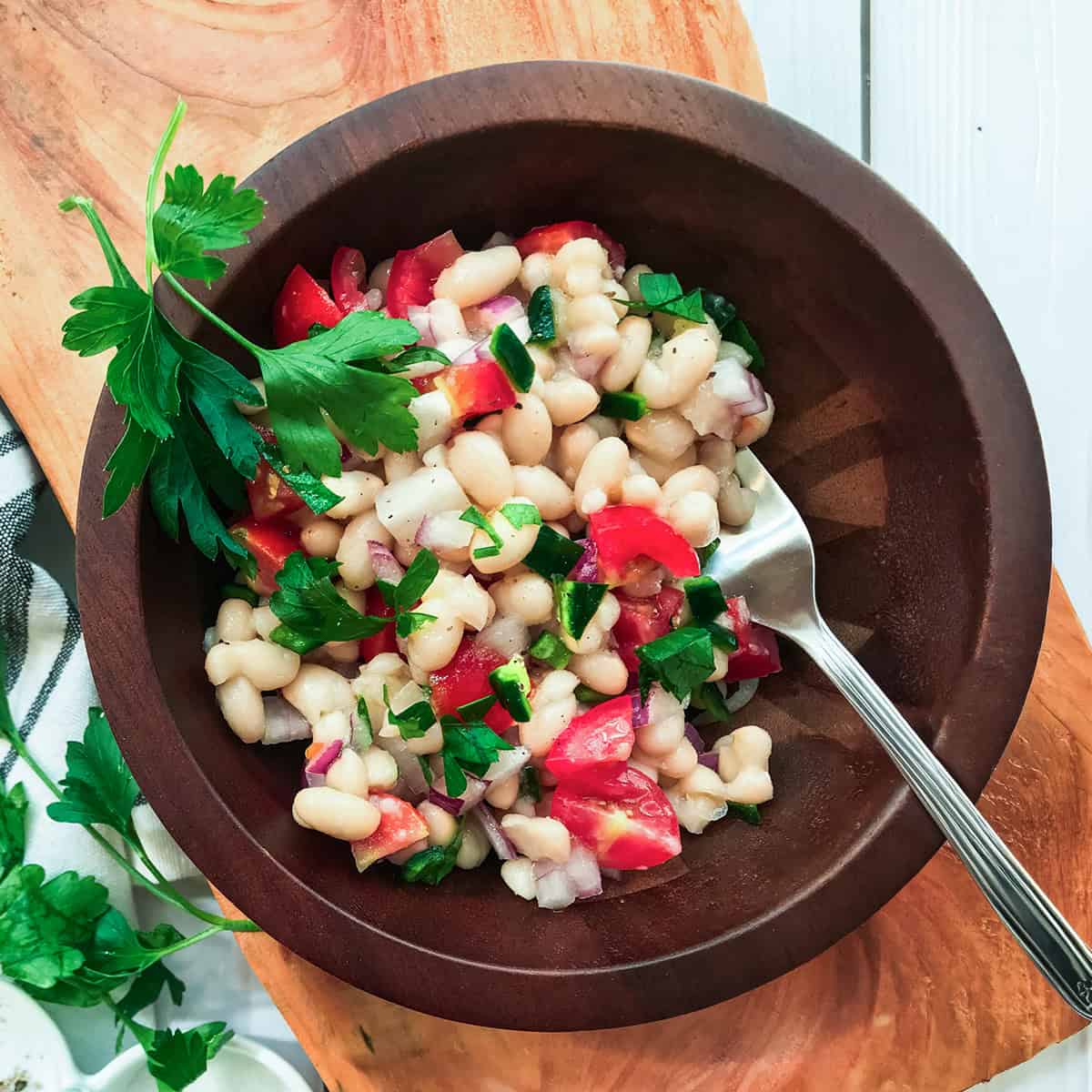 A wooden bowl filled with White Bean Salad, featuring white beans, chopped tomatoes, onions, parsley, and a metal spoon.