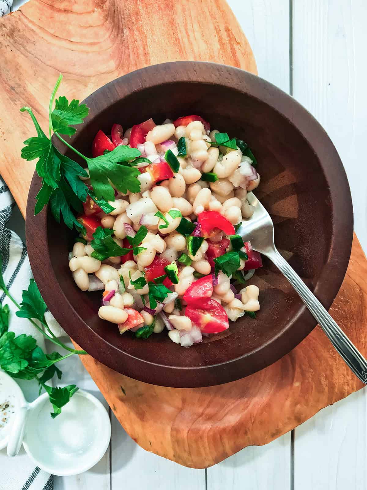 A wooden bowl of White Bean Salad with chopped tomatoes, red onion, and parsley, served with a fork on a wooden board.