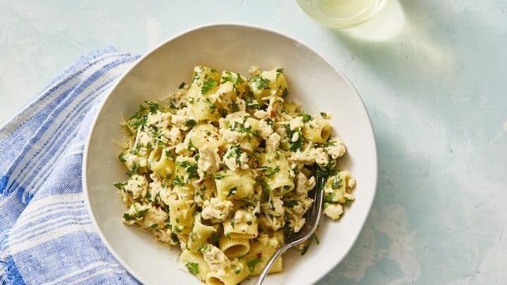 A bowl of rigatoni pasta with crumbled cheese and herbs, served with a fork beside a blue-striped napkin and a glass of white wine—perfect for enjoying during Labor Day Week.