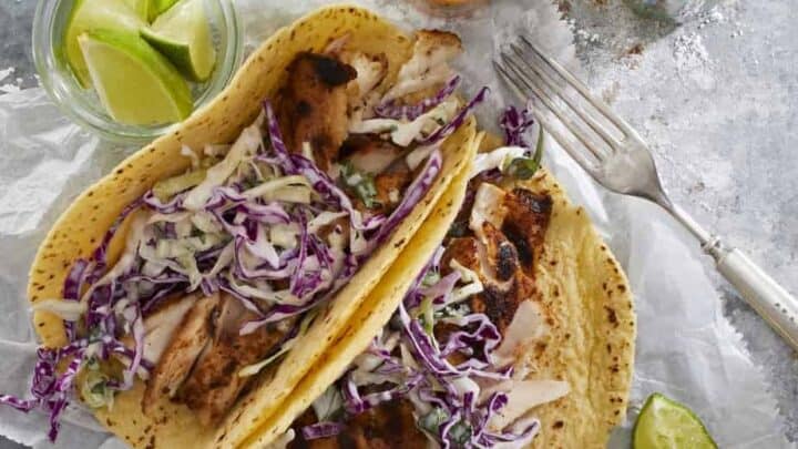 Perfect for early fall dinners, two grilled chicken tacos with shredded cabbage slaw on tortillas are served with lime wedges, hot sauce bottles, and a fork on a parchment-lined surface.