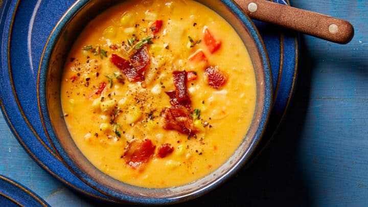 A bowl of creamy corn chowder topped with bacon bits, black pepper, and herbs, perfect for early fall dinners, served on a blue plate with a spoon.