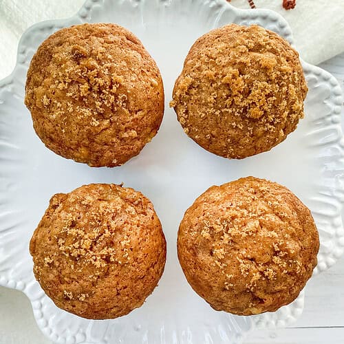 Four golden-brown banana muffins sit on a white, decorative plate. The plate is placed on a white surface with a striped cloth nearby. These freshly baked delights have a crumbly texture on top, perfect for those seeking the ultimate banana muffin recipe.