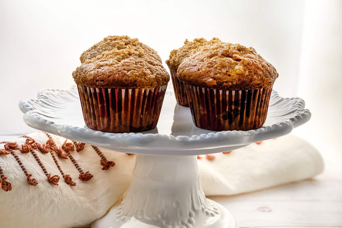 Three delightful banana muffins with crumbly toppings sit on a white, ornate cake stand. The softly lit background complements the scene, where a white napkin and subtle hints of brown tassels add a touch of elegance to this inviting recipe display.