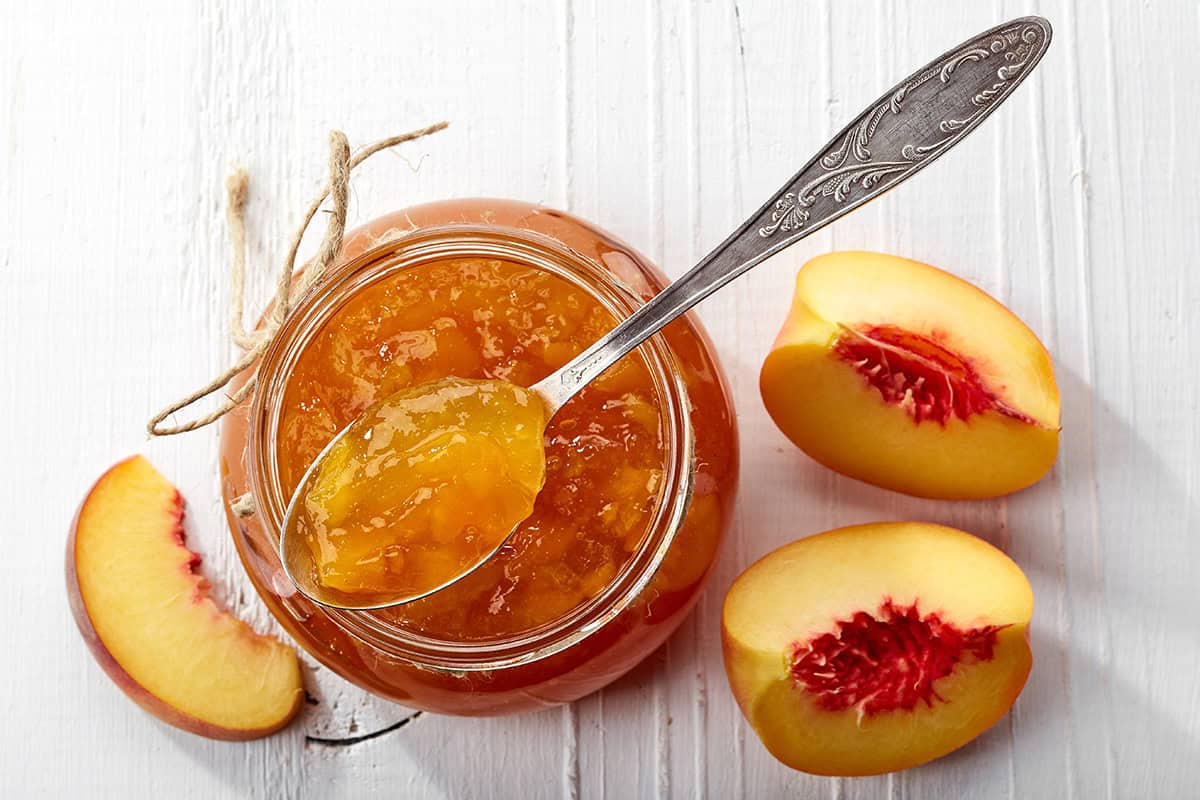 An open jar of Peach Jam with a spoon rests on a white table beside a whole peach and several peach slices.